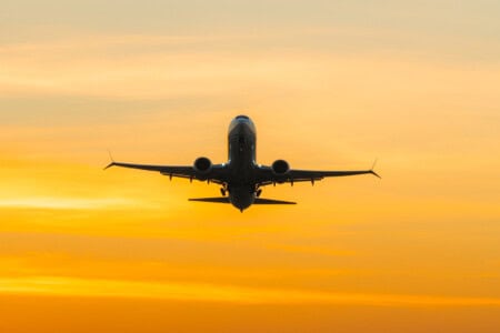 An airplane rises into the sky illuminated by a vibrant sunset in warm shades of yellow and orange